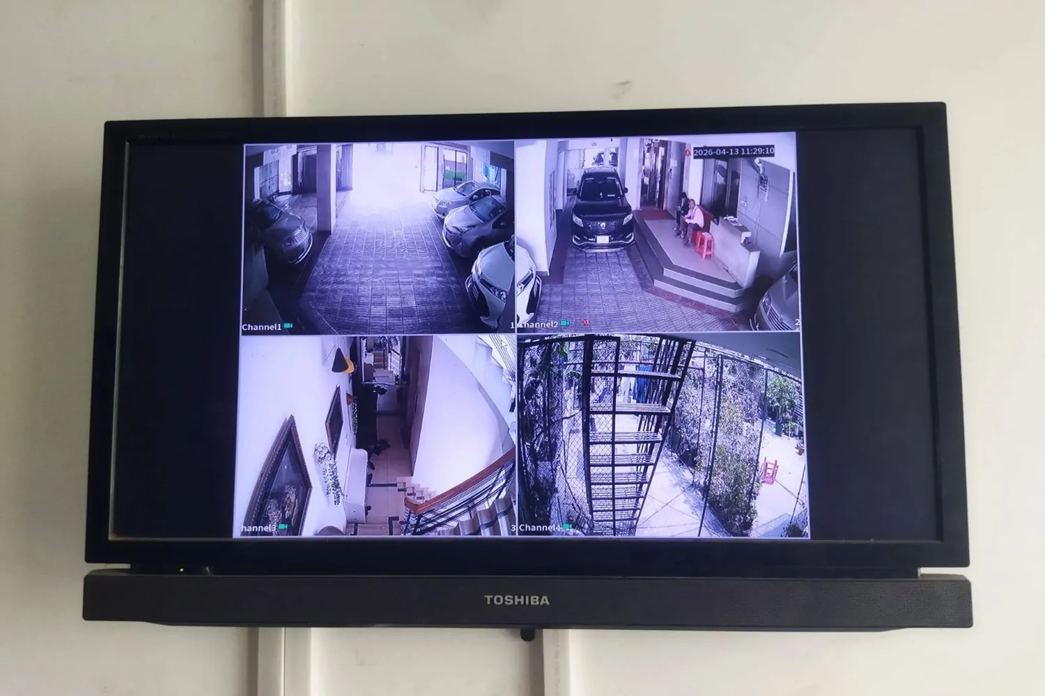 An IT technician installs a white dome camera next to a large monitor displaying a 4-channel split-screen security feed showing a parking lot, server room, and office hallways.