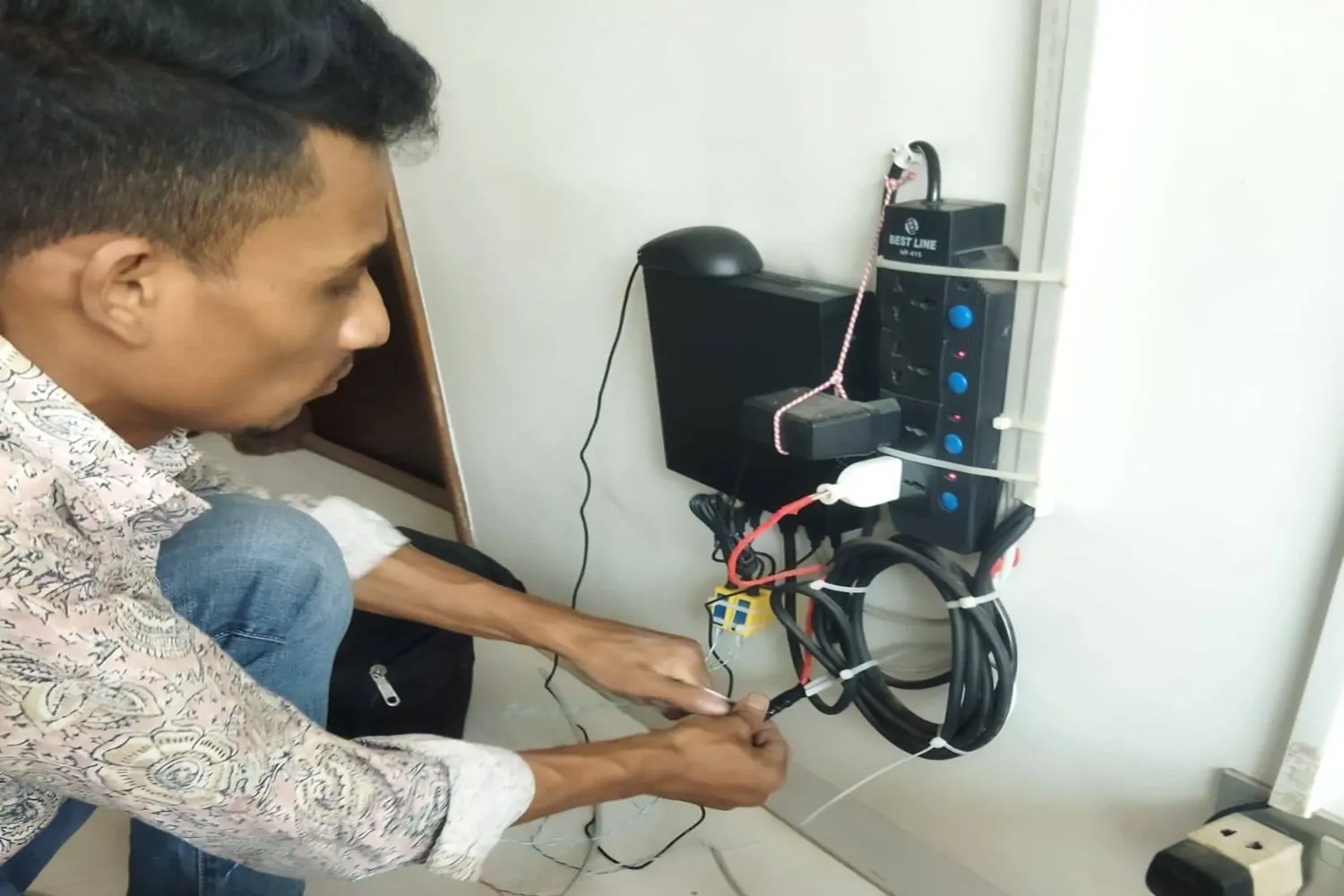 A technician in a patterned shirt and jeans working on a complex wall-mounted electrical and networking setup, organizing black cables and power strips.